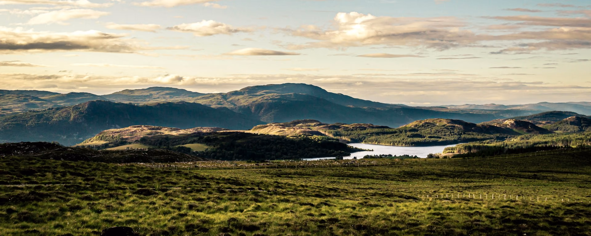 Highland loch and hills seen across a grassy foreground under broken cloud