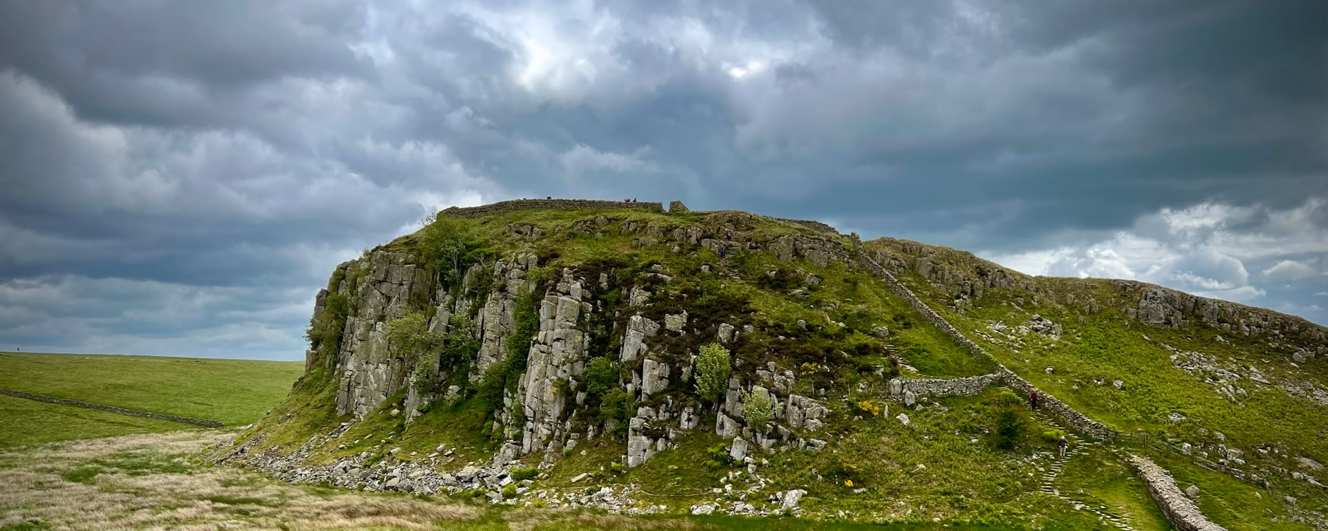 Paved path and stone wall climbing a crag under heavy cloud