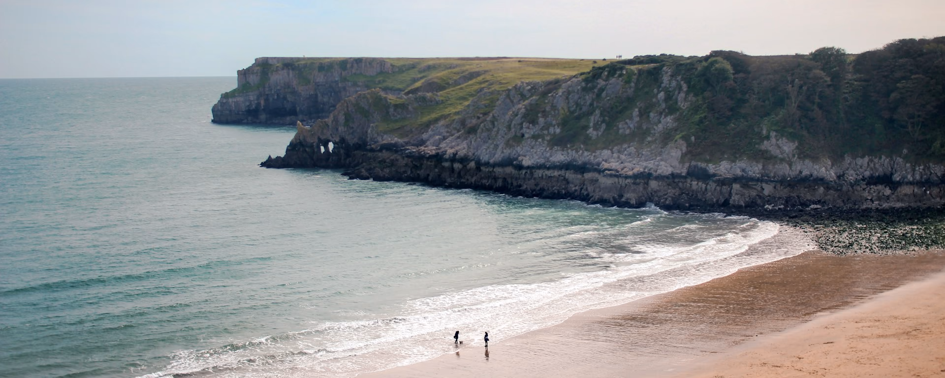 Two walkers on a sandy beach below grassy cliffs and a headland