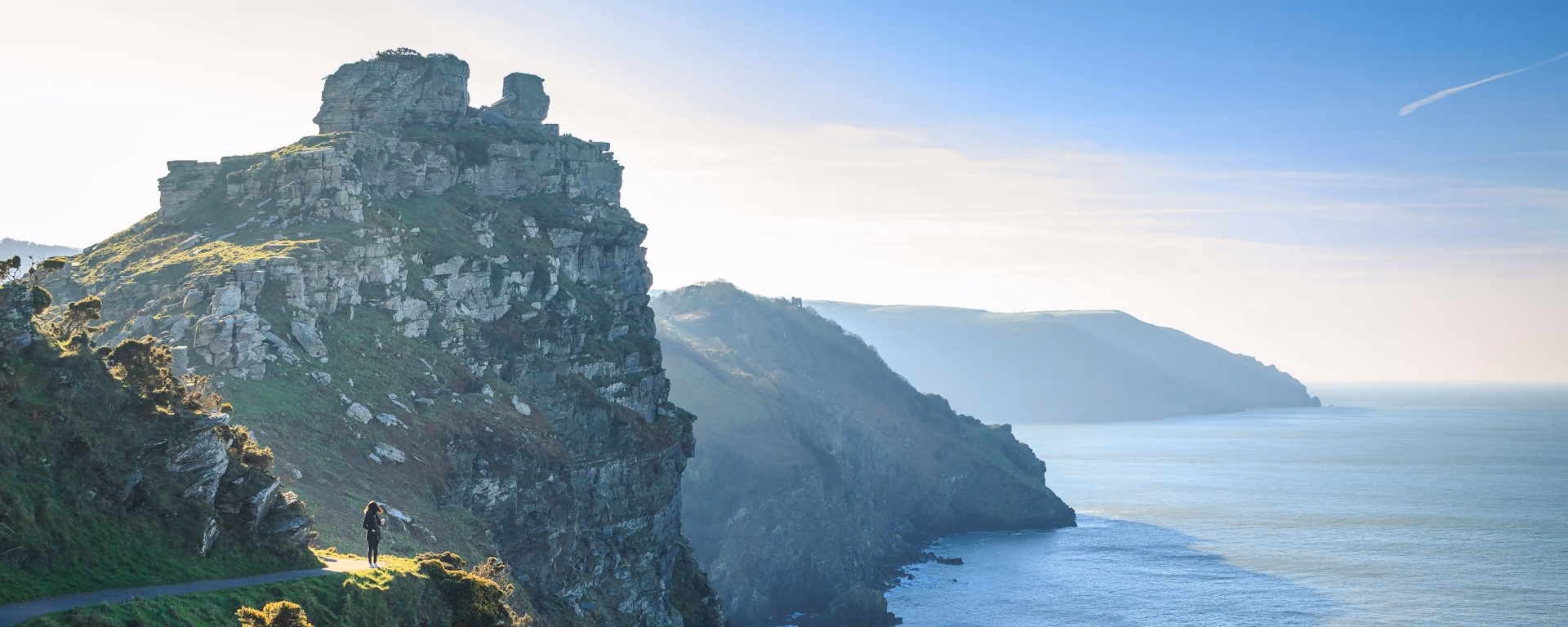 Coast path on a grassy clifftop, with a walker looking out over sea stacks and the sea