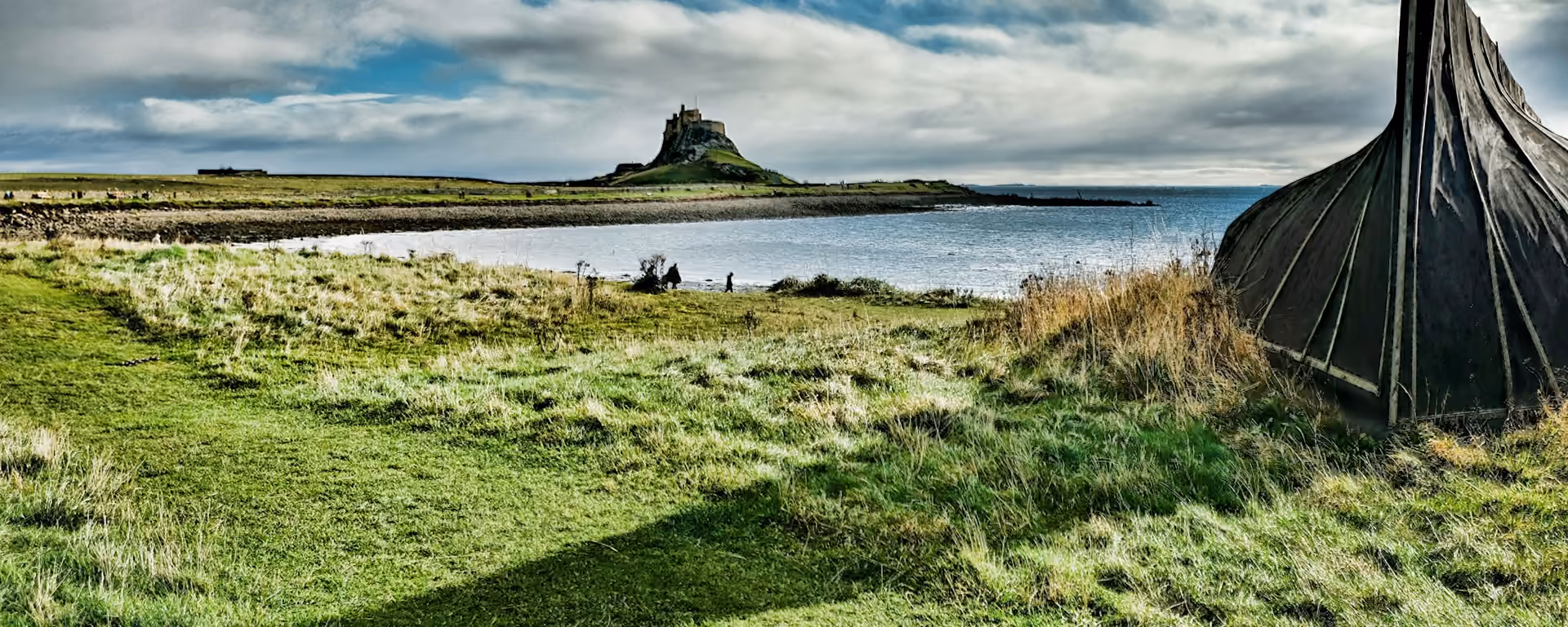 Lindisfarne Castle across a bay, from a grassy shore with an upturned boat